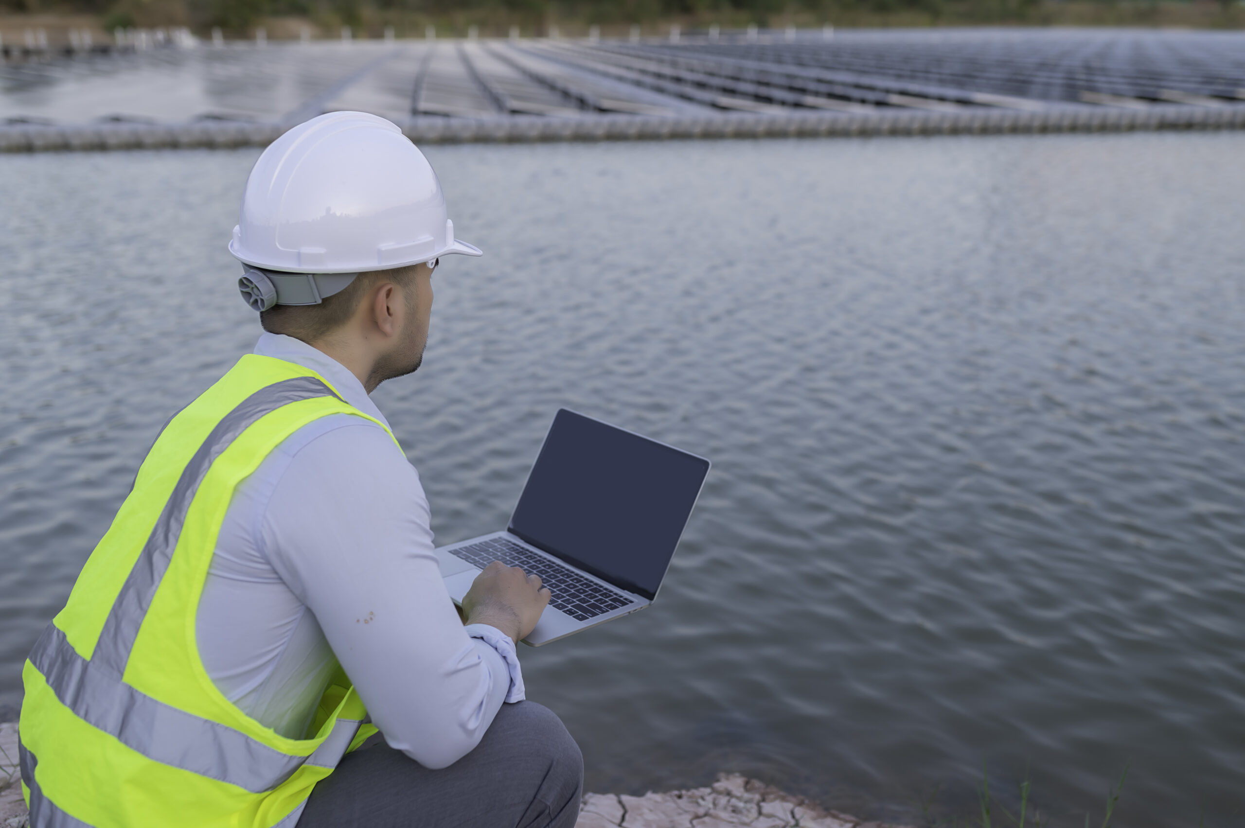 Engineer in a hard hat and safety vest using a laptop beside water